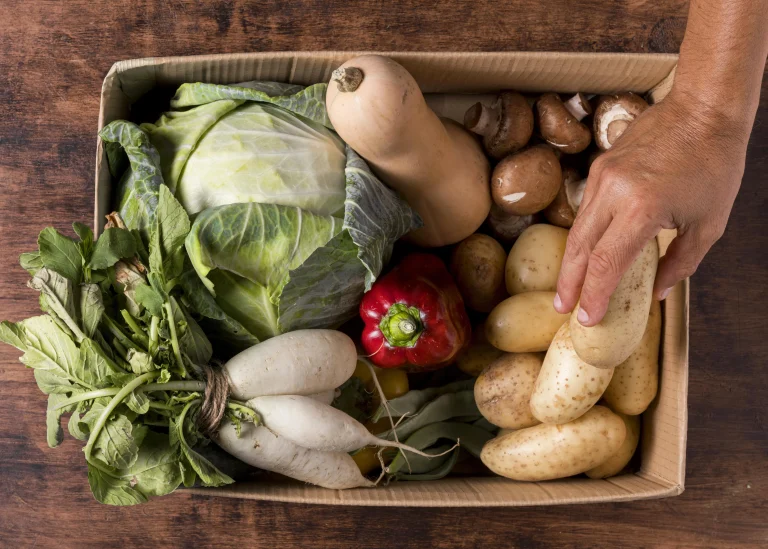 Pesticides free, Fresh organic vegetables including cabbage, daikon radish, potatoes, butternut squash, mushrooms, and bell pepper in a cardboard box on a rustic wood table, representing pesticide-free whole foods for autoimmune health.
